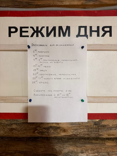 The image shows a handwritten schedule pinned to a white board with a red stripe at the top. The schedule is written in Russian. Large black letters at the top of the board spell out "РЕЖИМ ДНЯ," which translates to "Daily Schedule." Below this, a smaller heading reads "Распорядок дня осужденных," meaning "Schedule for the condemned."
The schedule details the daily routine:
* **6:00** - подъём (wake-up)
* **7:00** - ЗАВТРАК (breakfast)
* **7:30 - 8:00** - ПОСТРОЕНИЕ, ПЕРЕКЛИЧКА, РАЗВОД НА РАБОТУ (assembly, roll call, dispatch to work)
* **13:00 - 14:00** - ОБЕД (lunch)
* **19:00** - УЖИН (dinner)
* **20:00** - ПОСТРОЕНИЕ, ПЕРЕКЛИЧКА (assembly, roll call)
* **20:30 - 21:00** - ЛИЧНОЕ ВРЕМЯ ОСУЖДЕННОГО (personal time for the condemned)
* **22:00** - ОТБОЙ (lights out)
The schedule also includes weekend hours:
* **Суббота** - хоз. работы 1 час. (Saturday - 1 hour of housekeeping duties.)
* **Воскресенье** - с 15:00 до 16:00 - ползанятие. (Sunday - from 15:00 to 16:00 - half-session, likely referring to a period of activity or instruction.)
The overall environment appears to be utilitarian, with the wooden background suggesting an interior space that might be a facility or institution. The presence of pushpins indicates the schedule is intentionally displayed for viewing. The context implies this is a schedule within a correctional facility or a similar institution for individuals who are "condemned" or "convicted."
There are no people visible in the image. The scene is static. The location is specified as Ust'-Pyzas, Russia, which is a known location for correctional facilities.