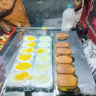 The image captures a street food stall, likely in Charfasson, Bangladesh, with a focus on a cooking station where egg-based items are being prepared. In the center, a metal griddle features multiple oval molds. On the left side of the griddle, several eggs are cooking; some show bright yellow yolks, while others appear to be just egg whites. Steam is faintly visible rising from the hot surface.

To the right of the griddle, a row of approximately seven prepared items resembling small, round buns or patties filled with what appears to be cooked egg are neatly lined up. These could be a type of egg burger or sandwich.

Various utensils and ingredients surround the cooking area. A red-handled tool and a wooden-handled brush are near the griddle. A red tray containing sliced green citrus fruits (lemons or limes) is visible on the far left. On the workstation, there's a small container with a yellow-green lid and a dark bottle.

In the background, a person, mostly obscured and wearing an orange/brown garment, is seen on the left. On the right, a child is partially visible, looking upwards with an open mouth, standing next to a large clear plastic bottle with a blue cap, containing a white liquid. Adjacent to the child and the bottle is a black battery connected with red and black wires, suggesting a makeshift power source for the stall. A white cloth with a patterned design is also visible on the lower right. The overall setting suggests a bustling, functional street food environment. No readable text is clearly visible on any items.