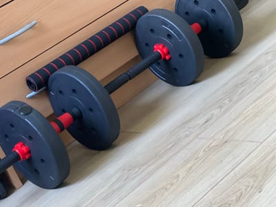 The image displays a set of adjustable dumbbells and a grip bar resting against a light-colored wooden surface, likely part of a wall or furniture. The dumbbells are dark gray with red collars and black threaded handles. One dumbbell is partially visible in the foreground on the left, and another is in the mid-ground, both with red star-shaped locking nuts. A third dumbbell is mostly visible in the background on the right. A black grip bar with red bands is positioned diagonally across the wooden surface. The floor beneath the equipment appears to be light wood laminate. There is no visible text in the image. The lighting suggests an indoor setting, possibly during the daytime due to the presence of light, but the specific time of day or weather conditions cannot be determined. The overall scene depicts exercise equipment, implying an activity related to physical fitness or strength training. No people are present in the image, and the emotions conveyed are neutral. The setting is domestic or a home gym environment.