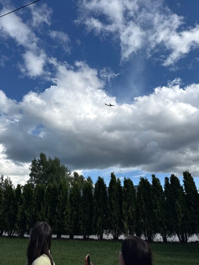 In an outdoor scene, likely in the city of Zhostovo, Russia, an airplane is visible in the distance against a dramatic sky filled with both bright white and dark gray clouds. The airplane is silhouetted against the lighter parts of the sky, appearing small in the frame. Below the sky, a dense line of tall, dark green coniferous trees forms a barrier across the middle ground. In the foreground, a grassy lawn is visible. Two individuals are partially seen in the lower portion of the image, facing away from the camera and looking upwards towards the sky. One person on the left has long, dark hair and is wearing a light-colored shirt. The other person on the right has dark hair and appears to be holding a mobile phone. The overall impression is a moment captured outdoors, possibly with people observing the airplane's flight.