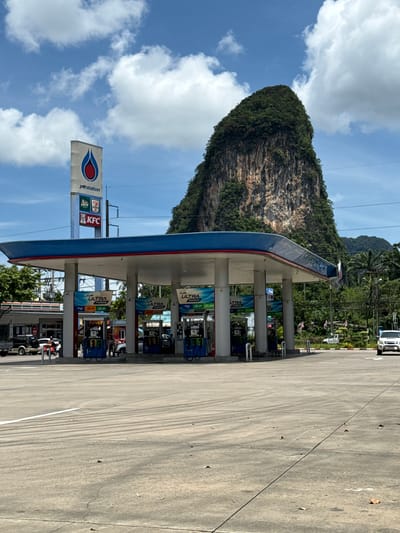 This is a street-level, eye-level, medium shot of a PTT gas station in Ao Luek, Thailand, with a dramatic, tree-covered limestone karst cliff in the background. The gas station has a blue and white canopy with red trim, supported by white pillars. Several gas pumps are visible under the canopy, with "PTT ULTRA FORCE" signage. A tall PTT sign is also visible, featuring the PTT logo along with logos for KFC and 7-Eleven. A white SUV is parked at the gas pumps on the right side of the frame. In the middle ground, there is a road with some vehicles, including a silver SUV and a white SUV, as well as a person standing near the gas pumps. The foreground consists of a concrete paved area with white road markings and scattered leaves. The sky is blue with scattered white clouds, and lush green trees and foliage are visible behind the gas station and around the base of the cliff. The limestone cliff is a dominant feature, with visible rock striations and dense vegetation covering its peak and upper slopes. The overall scene depicts a commercial area in a tropical environment.