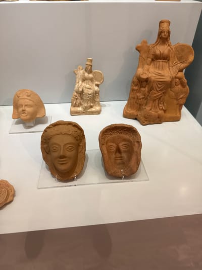 The image displays several ancient terracotta artifacts, likely from a museum exhibition. In the foreground, two terracotta heads are showcased on clear plastic stands. The head on the left is slightly angled, with simplified facial features and a round headdress. The head on the right is facing forward, also with stylized features and curly hair. Behind these, to the left, another terracotta head is visible, less detailed than the two in the foreground. In the middle ground, a small, light-colored terracotta figurine of a seated figure with a circular object in its hands stands on a pedestal. To the right, a larger, darker terracotta sculpture depicts a seated female figure with a headdress, a draped garment, and multiple figures clustered around her base. The objects are arranged on a white surface, with a light grey wall in the background. These artifacts are likely part of the collection at the Heraklion Archaeological Museum in Heraklion, Greece, known for its extensive Minoan and Hellenistic antiquities.