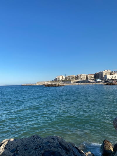The image shows a coastal view in Beau Fraisier, Algeria. The scene is divided between a clear blue sky and the turquoise water of the Mediterranean Sea. Along the coastline, there are several multi-story buildings, a promenade, and some trees. The shore is lined with rocks and some evidence of wave action is visible.