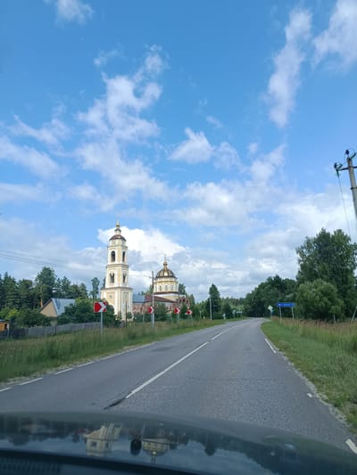 The image captures a road curving through a rural landscape under a bright, partly cloudy sky. On the left side of the road, a distinct church with a tall bell tower and a dome stands prominently, surrounded by trees and a wooden fence. A residential building is visible near the church. Several red and white directional signs are placed along the roadside, indicating curves in the road. On the right side, a utility pole with electrical insulators stands, and a blue road sign indicates "С. Покров 2,2," suggesting the village of Pokrov is 2.2 kilometers away. The foreground shows the reflection of the church and sky on the hood of a vehicle, indicating the image was taken from a car. The overall environment appears to be that of a tranquil countryside setting in Russia, potentially near the village of Gribchikha, given the context of the prompt.