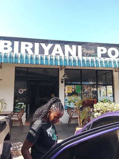 This image depicts the exterior of "Biriyani Pot," a restaurant located in Naivasha, Kenya, under a clear blue sky. The restaurant features a light-colored facade with a distinctive blue-green corrugated awning. Large white letters spell "BIRIYANI POT" above the entrance, with a smaller "Biriyani Pot Eat And Dream" logo positioned centrally. The entrance consists of double glass doors, one of which is ajar, revealing a dimly lit interior. Adjacent glass panels display promotional stickers, including "Halal," "Coffee," "Glovo," and "Uber Eats" logos. Outdoor seating comprising wicker-style chairs and small tables is arranged to both the left and right of the entrance, interspersed with potted plants, including a small palm and a flowering bush.

In the foreground, a dark-skinned female with braided hair, pulled back into a ponytail, stands facing downwards. She wears a dark t-shirt with a visible green graphic. Her posture suggests interaction with the interior of a partially visible purple vehicle, whose driver's side door is open on the right side of the frame. A portion of a tan/beige vehicle, including a tire, is visible on the far left. The ground is paved.