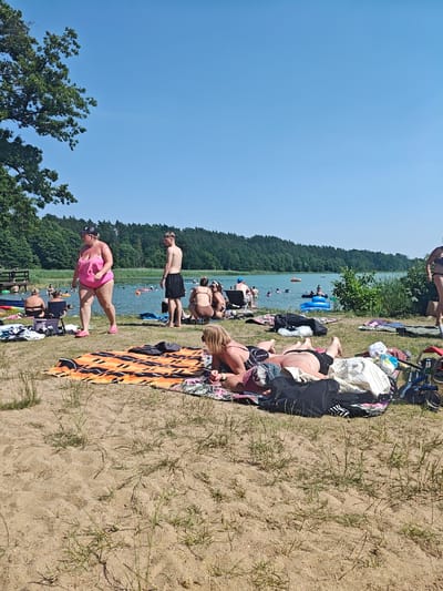 The image depicts a sunny day at a beach on Lake Piekary in Stare Miasto, Poland. The scene is populated with people enjoying recreational activities. In the foreground, several individuals are sunbathing on towels and blankets laid out on the sandy shore.
A woman in a bright pink one-piece swimsuit and pink flip-flops stands prominently in the mid-ground, facing away from the camera. Next to her, a man in black swim trunks stands shirtless.
The lake itself is visible in the background, with numerous people swimming, floating on inflatable toys, and wading in the water. A line of trees forms a dense backdrop along the far shore of the lake. The sky is a clear, bright blue. The overall impression is one of a typical summer day at a popular lakeside leisure spot.