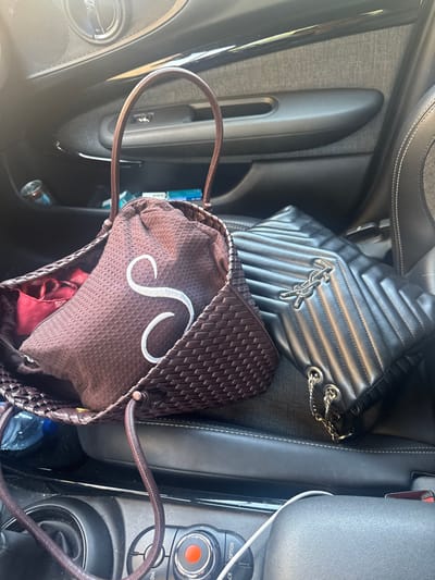 The following describes a scene observed within a vehicle located in Cidade da Maia, Portugal.
The image depicts the interior of a vehicle, specifically focusing on the passenger seat and the adjacent door panel and a section of the center console. No individuals are visible.
Two handbags rest on a dark-colored, possibly black leather, car seat. On the left, a dark brown handbag with a woven texture and a single, looped handle contains a lighter brown drawstring pouch that features a prominent white embroidered capital 'S'. A red fabric item is partially visible within this pouch. To the right, a black quilted leather handbag, bearing a silver-toned 'YSL' logo, lies on its side, revealing a silver metal chain strap.
The vehicle's interior elements include a dark gray door panel on the left, featuring a textured fabric insert, a door handle, and a window control button. A storage pocket within this door panel holds a blue Monster energy drink can and a blue and white cigarette pack. A portion of the center console is visible in the lower foreground, displaying a circular control dial with a red central indicator, and a white charging cable lying across it. The scene is illuminated by bright daylight, suggesting the image was captured during the day.