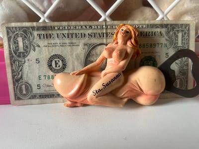 This media file features a close-up, eye-level shot of a novelty item placed in front of a U.S. one-dollar bill. The novelty item is a figurative sculpture of a nude woman with blonde hair, sitting astride a phallic-shaped object. The sculpture is predominantly a pale pinkish-peach color with some reddish-brown accents on the hair and lips. The phrase "Sta. Susanna" is written in black script on the thigh of the sculpture. Behind the sculpture and the dollar bill, a white, lattice-like structure is visible, suggesting a cage or fencing, and a soft, textured surface, possibly fabric or fur, is in the background. A metal bottle opener is partially visible on the right side of the frame.
The U.S. one-dollar bill displays the text "FEDERAL RESERVE NOTE," "THE UNITED STATES OF AMERICA," "THIS NOTE IS LEGAL TENDER FOR ALL DEBTS, PUBLIC AND PRIVATE," the serial number "8858977 N," and "WASHINGTON, D.C." Other visible text includes the denomination "5" printed multiple times, and the seal of the Federal Reserve Bank.
The lighting appears to be artificial and bright, casting minimal shadows. The setting seems to be indoors, on a white, textured surface, likely a table or shelf. There are no obvious cues indicating a specific time of day or weather conditions. The overall impression is a staged, still-life composition focusing on the novelty item and the dollar bill.