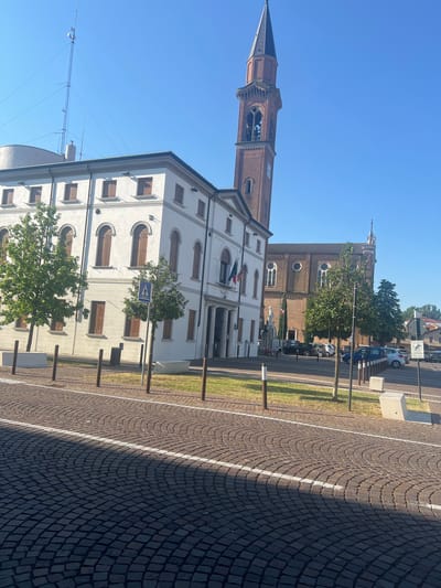 The image shows a view of a town square in Campodarsego, Italy, on a clear, sunny day. In the foreground, a cobblestone street is visible, with a white painted line marking a border. Beyond the street, there is a white building with brown shutters on its windows. To the right of this building, a tall, brick bell tower rises into the blue sky. Further in the background, a brick church with arched windows is visible. Several trees are planted in planters and along the sidewalk. There are a few cars parked on the street. A pedestrian crossing sign is visible on the left side of the image. The overall scene depicts a quiet, orderly urban environment.