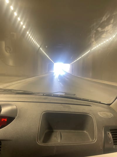 The image depicts the interior view from a vehicle driving through a well-lit tunnel in Amman, Jordan. The perspective is from the driver's or front passenger's seat, looking forward through the windshield.
The tunnel appears to be constructed of plain, light-colored concrete walls and ceiling. Both upper corners of the tunnel are lined with continuous rows of bright, white LED-style lights, providing illumination. The asphalt road surface within the tunnel is visible and appears to be in good condition. Ahead, a distant and intensely bright light source indicates the tunnel exit, creating significant glare and lens flare on the vehicle's windshield. This light suggests bright daylight outside.
In the foreground, the dark gray dashboard of the vehicle occupies the lower portion of the frame. The windshield wipers are visible resting horizontally at the base of the windshield. The windshield itself appears dusty or smudged, contributing to the light diffusion. On the left side of the dashboard, a small red digital display shows "12:46". Centered on the dashboard is an open, rectangular storage cubby. To the right, a portion of an air vent grille is visible. No individuals are depicted, but the scene implies movement within the vehicle.