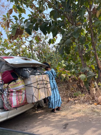 The scene, located in Nandu, Nigeria, depicts an outdoor environment under clear daylight. A light-colored vehicle, possibly an SUV or minivan, is parked on a dirt shoulder next to a dark road surface. Its rear tailgate is open, revealing a heavily packed cargo area. Visible items include a large, rectangular cardboard box wrapped in clear plastic and secured by a black elastic cargo net with additional yellow and red ropes. To its left, a red backpack and a multi-colored striped woven bag are also present, alongside other dark, bundled packages. A single individual, viewed from the back, is seen leaning towards the loaded vehicle. This person is wearing a long, light blue dress with a dark patterned design and dark flat shoes. The surrounding area is characterized by dense green foliage, comprising trees with broad leaves and dry, sparse undergrowth on the ground.