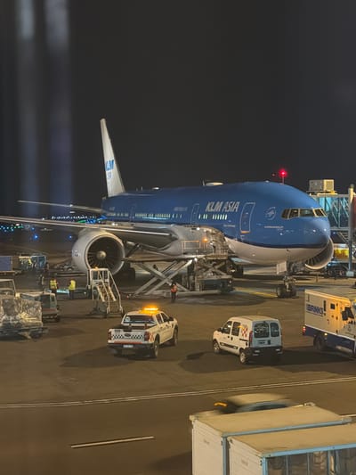 At Aeropuerto Internacional Ezeiza, Argentina, a KLM Asia Boeing 777 aircraft is parked on the tarmac at night. The aircraft is predominantly blue with white accents and features the KLM logo on its tail. Various ground support equipment, including baggage carts and a service vehicle with orange flashing lights, surround the aircraft. Several individuals in high-visibility vests are visible near the aircraft, engaged in ground operations. A Brinks armored truck is also present on the tarmac. The airport environment is illuminated by artificial lighting, and the sky is dark.
