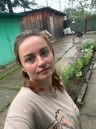 The image depicts a selfie taken outdoors, presumably in a yard or rural setting. A woman with light brown hair and green eyes is the central subject, looking directly at the camera. She is wearing a light brown t-shirt with a graphic print on the front, and there are some white paint-like splatters on the left shoulder area.

In the background, there is a weathered wooden structure, possibly a shed or outbuilding, with vertical wooden planks. To the left of this structure is a bright green door. A paved pathway runs through the scene, and along the right side of the pathway is a flower bed with various colorful flowers, including purple, pink, and white varieties, as well as green foliage.

Further in the background, a wooden fence with green paint encloses the area. A dog, appearing to be a medium-sized breed with a mix of brown and black fur and a bushy tail, is visible in the middle ground, walking on the wet pavement. Behind the dog, a white rectangular object, possibly a piece of discarded appliance, rests on the ground. The ground appears wet, suggesting recent rain. Overhead, tree branches and foliage are visible, with a cloudy sky in the distance. The overall impression is a candid shot taken in a somewhat unkempt, possibly rustic environment.
