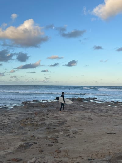 A surfer stands on the rocky shoreline of Thirroul Beach in Australia, holding a surfboard. The beach stretches towards the ocean, with waves breaking near the shore. The sky is a mix of blue and light orange with a small moon. Clouds are scattered throughout the sky.