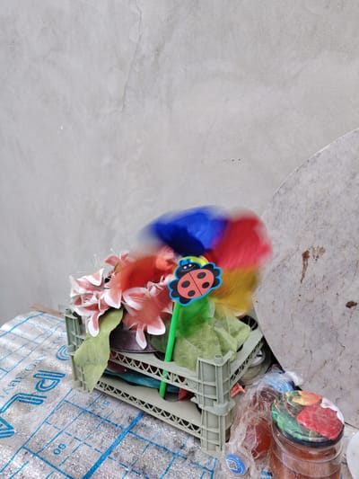 A plastic crate filled with artificial flowers and a multi-colored fan or blur object is placed on a table covered with a patterned sheet. A ladybug decoration is attached to a green stick protruding from the crate. In the background, a rough, gray concrete wall serves as the backdrop. To the right of the crate, a large, round, light-colored object, possibly a stone or marble slab, leans against the wall. A glass jar filled with a brown substance, possibly jam or honey, sits on the table to the right of the crate. The scene is likely an outdoor setting, perhaps a yard or street in the city of Yangiiul, Uzbekistan, given the style of construction and materials visible.