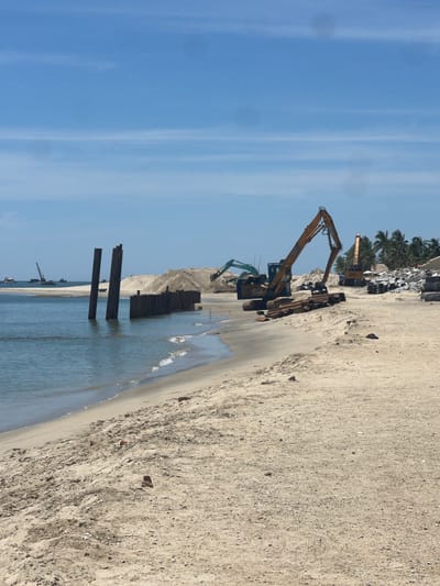 The image depicts a beach scene in Hoi An, Vietnam, with construction activities underway. Two yellow excavators are prominent subjects, positioned on the sandy shore. One excavator is in the foreground, its arm extended upwards, while another, slightly behind it, is also engaged in work. A smaller, green excavator is partially visible in the lower left quadrant, suggesting ongoing earthmoving.

The setting is a beach with a clear blue sky and calm water. The sand appears dry and coarse, with some debris scattered across it. In the background, a line of palm trees suggests a tropical or subtropical location. Further in the distance, there are mounds of sand or earth, possibly created by the construction efforts, and piles of what appears to be rubble or building materials. Several dark, rectangular structures, possibly pilings, are erected in the shallow water near the shore, indicating the construction of a sea defense or pier.

The activity taking place is clearly construction, likely related to coastal development or reinforcement. The time of day appears to be daytime, given the bright sunlight and clear sky, though the exact time is not specified. The weather seems fair. There is no discernible text in the image. The overall impression is one of industrial activity impacting a natural beach environment.