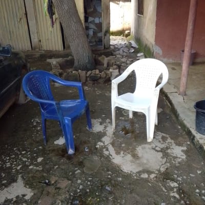 The image depicts an outdoor scene, likely in Mangu, Nigeria, with two plastic chairs as the main subjects. A blue chair is positioned on the left, and a white chair is on the right, both placed on a wet, uneven ground scattered with rocks and debris. In the background, a tree with a textured trunk stands prominently, with its branches obscured. To the left of the tree, a corrugated metal structure with some hanging laundry suggests a dwelling or storage area. Further back, an open doorway reveals a wall adorned with mosaic tiles, hinting at an interior space. The overall setting appears to be a utilitarian or somewhat unkempt outdoor area, possibly a yard or an alleyway. The ground's wetness suggests recent rain or a damp environment, and the lighting indicates it's daytime, possibly overcast. No people or animals are visible, and there is no visible text in the image.