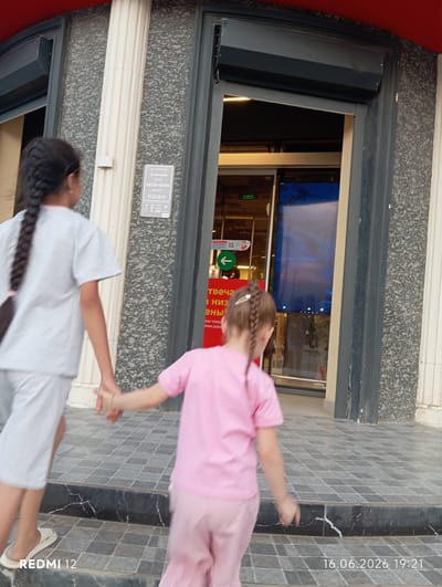 Two young girls are walking away from the camera towards the entrance of a building. The girl on the left is wearing a light gray tracksuit and has her hair in a braid. The girl on the right is wearing a pink t-shirt and pink pants, and also has her hair in a braid. They are holding hands. The building entrance appears to be a store or business, with a sign on the wall to the left of the entrance displaying operating hours. A digital screen is visible inside the entrance, displaying what appears to be a sky or water scene. The ground in front of the entrance is paved with gray square tiles. The timestamp on the bottom right of the image indicates the date is June 16, 2026, at 7:21 PM. The location is identified as Bekabad, Uzbekistan.