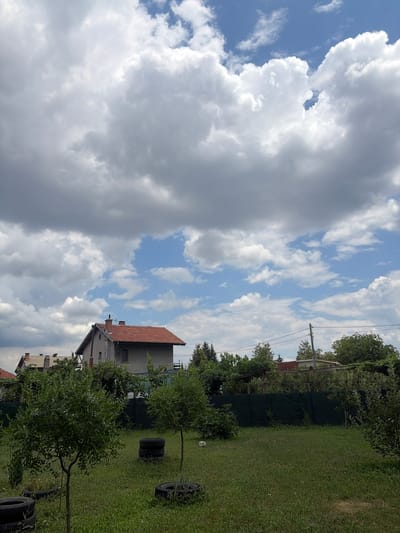 In the city of Yulievo, Bulgaria, a bright, cloudy sky looms over a residential area. In the mid-ground, a two-story house with a red tile roof and a prominent chimney is visible, flanked by trees and foliage. To the left, a section of another house with a similar red roof is peeking out. The foreground features a grassy yard with scattered tires, some stacked, and two small trees. A power line and pole are visible in the background, adding to the suburban feel of the scene.