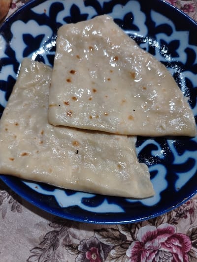 Three flatbreads rest on a blue and white patterned plate. The bread is a light beige color, marked with small brown spots and a glistening sheen suggesting an oil or butter glaze. The plate features a symmetrical, stylized floral or leaf design in white against a dark blue background. The plate sits on a table covered with a floral-patterned tablecloth in muted tones of pink, brown, and beige.