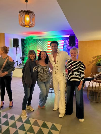A group of five people pose in a room with a stage backdrop featuring an American flag and a peacock feather design, possibly after a performance or event. One individual is dressed in a white Elvis Presley-style jumpsuit. There's a light fixture on the wall, speakers set up near the background, and decorative floor tiling. The location is Grimaud, France.