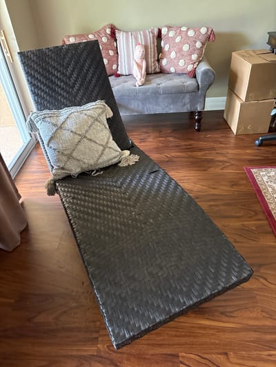 This is a top-down, eye-level shot of an indoor scene, likely a living room or lounge area, with a focus on furniture. The floor is made of dark, polished wood planks with a distinct grain pattern.
On the left side of the frame, there's a light-colored wall. Leaning against it is a gray upholstered loveseat or chaise lounge with tufted details and a carved wooden leg. Several decorative pillows are stacked on the loveseat. The bottom pillow is pink with white polka dots and decorative tassels. Above that is a rectangular pillow with a striped pattern in white and reddish-pink, and a light pink plush toy or pillow with a duck face.
In the center and right of the frame, a large, dark brown woven rectangular ottoman or coffee table is positioned diagonally. It has a textured, basket-weave pattern. Resting on the ottoman is a square decorative pillow with a gray textured fabric and a diamond-patterned stitch. This pillow also has tassels on its corners.
In the upper left portion of the image, two cardboard boxes are visible, stacked on top of each other. One box has handwritten text that appears to say "Blink" and "John."
Further to the right, a small portion of a rug with a traditional pattern and a red border is visible on the wooden floor. The scene appears to be lit by natural light, suggesting daytime. The overall impression is one of a lived-in, comfortable space, possibly in the process of being organized or decorated. There are no people visible in the image.