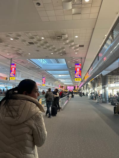 A long hallway is filled with travelers on a moving walkway in an airport. Signs for gate B32 hang above the walkway. The hallway features a white tiled wall with recessed circular shapes. A woman in a light-colored, puffy jacket is looking towards the end of the walkway. Other people are seen traveling along the walkway, some with luggage. Bright lighting illuminates the scene.