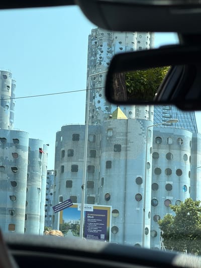 The image shows a view from inside a vehicle looking at the exterior of a cluster of cylindrical buildings with rounded-square and circular windows. One building has a red flag protruding from an opening. A billboard for "GEDIM" and "EKLO" is visible between two of the buildings. The billboard advertises a new residential complex and mentions dates in June and the city of Nanterre. The sky is clear and blue, and green trees are visible in the foreground and background, as well as a reflection of trees and a tree trunk in the car's rearview mirror. The buildings are light blue or gray and appear to be made of concrete or a similar material, with a patterned facade of lighter and darker shades and small, square or circular openings.