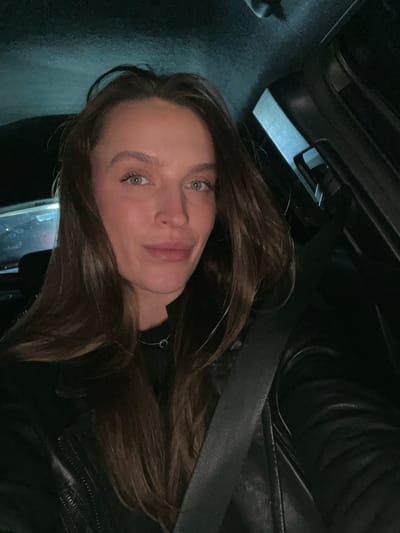 The image displays a portrait of a young woman with long, dark brown hair, fair skin, and striking green eyes, looking directly at the camera with a gentle smile. She is wearing a dark, possibly leather, jacket or top, and a thin silver necklace with a small circular pendant is visible around her neck. A black seatbelt is diagonally stretched across her chest and shoulder, indicating she is seated inside a vehicle.
The setting appears to be the interior of a car, likely at night or in low-light conditions, as the background is predominantly dark and indistinct, showing only vague shapes of the vehicle's interior. The lighting primarily illuminates her face, creating a soft glow. There are no specific visual cues within the image to identify the precise location as Braćeni, Montenegro, other than the general context of being in a car. No text is visible. The mood conveyed is calm and composed.
