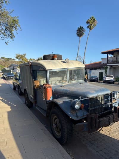 The image captures a static daytime street scene in Malibu, United States, under a clear blue sky. A heavily weathered, faded blue-gray vintage utility truck, consistent with a military-style vehicle, is parked curbside on the right, facing towards the viewer's left. The truck displays extensive surface rust and paint degradation. It features a box-style utility body, a split windshield, round headlights, an amber turn signal, and a square grille with a robust, rusted front bumper incorporating chain links. An orange, corroded jerry can is suspended by a chain from the driver's side, positioned below an external, vertical exhaust pipe. A spare tire and an amber rotating beacon light are mounted on the cab roof. The vehicle's large, aggressive-tread tires rest on a light-colored sidewalk composed of rectangular pavers, which is partially shadowed.
Behind the utility truck, a dark SUV and a dark sedan are parked along the same curb. Across the street, a two-story building exhibiting Mediterranean-style architecture, characterized by terra cotta roof tiles, stucco walls, and a visible balcony with a wrought-iron railing, is present. In front of this building, a dark sedan and a white SUV are parked. Two tall palm trees are prominent against the bright sky on the right side of the frame. A green, tree-lined hillside with distant residential structures is visible on the left side of the background. No people or dynamic actions are depicted in the scene.