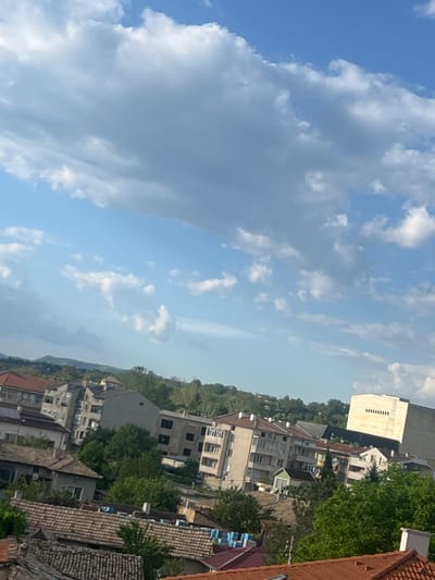 The image shows a view from above, possibly a rooftop, overlooking a cityscape on a partly cloudy day. The buildings appear to be residential and commercial, with a mix of architectural styles and some greenery interspersed. The sky is blue with white and gray clouds, suggesting fair weather. The location is indicated as Novi Pazar, Bulgaria.