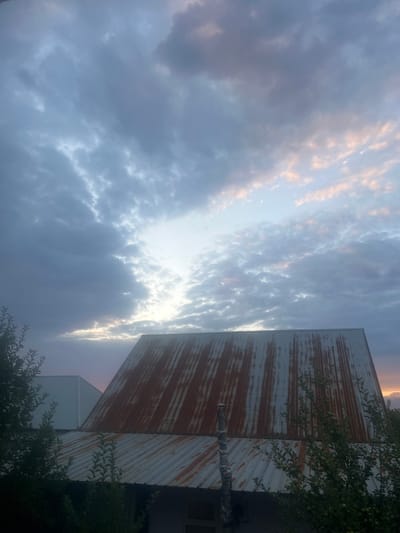 A low-angle, exterior, medium shot captures a rustic scene in Qaskeleñ, Kazakhstan, at dusk. The sky is filled with a dramatic array of clouds, ranging from dark gray to light pink, illuminated by the setting sun peeking through the gaps. In the foreground, two corrugated metal roofs of buildings are visible, one prominently featuring a weathered, rusty surface with streaks of orange and brown. The other roof, further back and to the left, appears lighter in color and possibly newer. Lush green foliage, possibly trees or bushes, partially obscures the lower parts of the buildings on the left and right sides of the frame. A pipe, possibly a chimney or vent, rises from the rusty roof. The overall impression is one of rural simplicity under a vibrant twilight sky.
