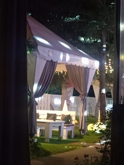 The image depicts an outdoor scene at night, likely at a venue in Bosteri, Kyrgyzstan. The foreground is dominated by a large, light-colored tent structure with decorative ruffled edges. The tent is illuminated from within by several bright lights, casting a warm glow and creating visible patterns on its surface. Draped fabric hangs from the tent, adding to its decorative appearance.

Behind the tent, a path or walkway is visible, bordered by a neatly mown lawn. In the background, there are structures that appear to be small, open-air seating areas or booths, with light-colored wooden walls and light purple tables and benches. Some of these structures are decorated with white horizontally arranged planks. String lights are visible in the upper background, suggesting a festive or ambient atmosphere. The overall impression is that of an outdoor event space or a venue set up for an evening gathering, possibly a restaurant or entertainment area. The time of day is clearly night, and there are no discernible weather cues. The scene evokes a sense of calm and festive ambiance. Visible text is minimal and partially obscured, but a circular logo that resembles an Instagram icon and some Cyrillic text can be faintly seen on one of the background structures.