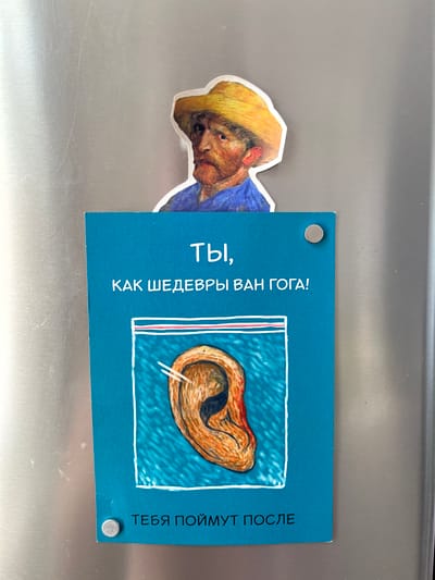 The media file shows a magnet on a refrigerator door. The magnet features a drawing of a person's head, appearing to be Vincent Van Gogh, with a yellow hat and blue shirt, on the left side. To the right of this drawing is a blue rectangle with text and an image. The text on the blue rectangle, written vertically in Russian, reads "ТЫ, КАК ШЕДЕВРЫ ВАН ГОГА!" on the left side of the image and "ТЕБЯ ПОЙМУТ ПОСЛЕ" on the right side. The image on the blue rectangle depicts a coiled ear inside a clear plastic bag, drawn in a style reminiscent of Van Gogh's artwork. The overall scene appears to be domestic, with the refrigerator as the setting. The lighting is consistent with indoor lighting. There are no obvious indicators of time of day or weather. The location context provided is Балаклавский округ, Ukraine.