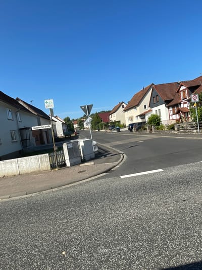 The image shows a residential street in Wartenberg, Germany, on a clear, sunny day. The street is paved and curves to the right. On the left side of the street, there is a sidewalk with a low concrete barrier and a wooden fence. A street sign reading "Lauterbacher Straße" is visible. Several houses with tiled roofs and light-colored facades are lined up along the street. A speed limit sign indicating "30 ZONE" is posted on the right side of the road. A traffic sign for cyclists is also present. Two dark-colored cars are parked in front of the houses. The sky is bright blue.