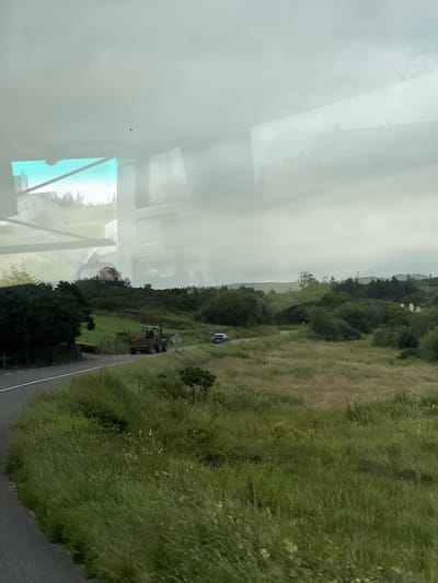 The image shows a rural landscape with a winding road in Falcarragh, Ireland. A green tractor with a trailer is driving on the road, followed by a dark-colored car. The landscape is characterized by rolling hills covered in lush green grass and scattered trees. The sky is overcast with clouds, suggesting a typical Irish day. Reflections of a building and the sky are visible on the surface through which the photo was taken, likely a window.