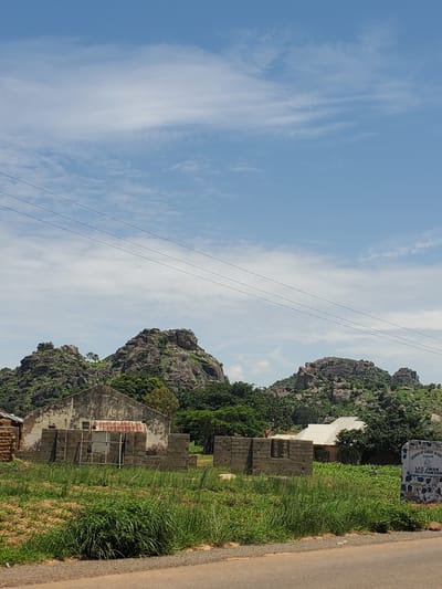 The image depicts a rural scene in Panyam, Nigeria, characterized by rocky hills in the background. In the foreground, there is an unfinished brick structure, suggesting ongoing construction or a derelict building. A partially visible house with a rusty corrugated iron roof stands to the left of the unfinished structure. To the right, a white roofed building is partially obscured by vegetation. A stone monument or signpost is also visible on the right side of the image, with text that appears to read "CHURCH OF CHRIST IN NIGERIA COCHIN LCC Jwak RCC Panyam." The environment is overgrown with grass and foliage, and power lines stretch across the sky. The sky is partly cloudy.