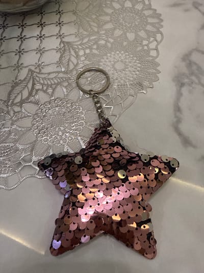 A sequined star-shaped keychain with a silver ring and chain is pictured. The keychain is lying on a white marble surface, partially on top of a silver lace placemat. The placemat has a floral design. The lighting in the image is a bit dim, creating some glare on the sequins. The image was taken at an angle, looking down at the keychain and placemat. The location is in the City of Pematang Siantar, Indonesia.