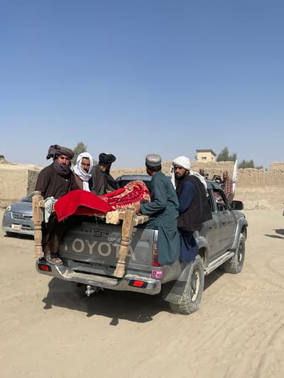 The image depicts an outdoor scene in Spin Boldak, Afghanistan, under a clear blue sky. Six men are gathered around the rear of a dark grey Toyota pickup truck, which has "TOYOTA" clearly embossed on its tailgate and a visible tow hitch.
A simple, unvarnished wooden cot-like frame, approximately 1.5 meters long, is positioned in the truck bed, extending slightly beyond the tailgate. This frame is largely covered by a prominent red fabric, patterned with dark designs, suggesting a rug or blanket.
All six men are dressed in traditional Afghan attire, including turbans or head coverings in white, black, and dark brown, along with long tunics and vests.
* The man on the far left, wearing a dark brown tunic and turban, is seated on the tailgate, his feet in sandals resting on the dirt ground, and his hands holding one end of the wooden frame.
* Another man, wearing a dark green tunic and a light-colored cap, is positioned inside the truck bed, near the center of the wooden frame, facing towards the front of the truck.
* A third man, in a dark blue tunic, black vest, and white turban, stands to the immediate right of the truck bed, appearing to interact with the frame.
* Three additional men are visible: one in a dark brown tunic and dark turban stands behind the man on the tailgate, another is partially visible further back in the truck's cab area, and a man in a light-colored tunic and white turban is on the far right, partially obscured.
The ground is unpaved, consisting of sand and dirt with visible tire tracks. In the background, characteristic mud-brick or earthen walls and structures of an arid region are visible. A light-colored sedan with a partial license plate "003" is parked behind the pickup truck on the left. On the right side of the image, a white flag with black calligraphy, consistent with a Taliban flag, is mounted on a pole near a building. A small, light-colored building with a dark roof and some sparse trees are also visible in the mid-ground.