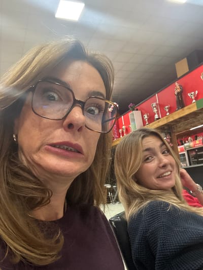 The image captures an indoor scene in Décines-Charpieu, France, featuring two females. In the immediate foreground, a female with light brown hair and prominent, dark-framed eyeglasses exhibits an exaggerated facial expression, characterized by wide eyes, a crinkled nose, and a slightly open mouth revealing visible dental aligners or braces on the lower arch. She wears a dark purple long-sleeved garment and a small hoop earring in her left ear. Behind her and to the right, a second, younger female with long blonde hair and a dark blue textured sweater smiles softly, looking towards the camera. Her left hand is positioned near her neck.
The background consists of a red wall with a horizontal green stripe at the top. A wooden shelf on the red wall displays several silver-colored trophies of varying sizes and a single brown religious figurine. Below the shelf, portions of a commercial refrigeration unit or display case are visible. The ceiling is a grid of light-colored square tiles, featuring recessed square fluorescent light fixtures. The environment suggests a commercial establishment, potentially a casual restaurant, bar, or community hall. The exact address within Décines-Charpieu is not discernible.