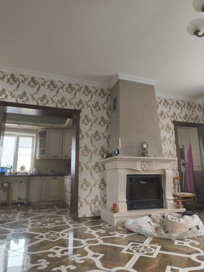 The image depicts an interior scene of a living room and adjacent kitchen. In the foreground, a cream-colored fireplace with decorative molding is visible, with a black insert for a fire. On the mantelpiece, there are two candlesticks with red candles, a decorative clock, and a small figurine of a mushroom. A patterned floor with brown and white tiles extends throughout the visible area. To the right of the fireplace, a wooden chair with a patterned seat and a pink towel hanging on its back is placed. A large beanbag chair with a floral pattern is situated in front of the fireplace.

To the left of the fireplace, an open doorway leads to a kitchen area. The kitchen features cream-colored cabinets, a countertop, a sink, and a window with white blinds. On the countertop, there are bottles of water, a plant in a pot, and other kitchen items. A radiator is visible beneath the window. The walls are adorned with ornate wallpaper featuring a repeating pattern of gold and cream foliage. The ceiling is white and features decorative molding.

The scene is set in a residential interior. Based on the metadata provided, the location is Мещерино, Russia.