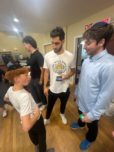 This image depicts an indoor scene involving multiple individuals within what appears to be a synagogue or Jewish community center in Bergenfield, United States.
In the foreground, three males are present. On the left, a young boy with reddish-brown hair and a black kippah, wearing a white t-shirt and black pants, is shown from the waist up, leaning slightly backward, with his mouth open, seemingly speaking or reacting. In the center, a young adult male with dark, curly hair and a short beard, dressed in a white t-shirt with a "Vans OFF THE WALL SINCE 1966" graphic and black athletic pants, holds a dark smartphone horizontally in his right hand, looking down at it. To his right, a second young adult male with dark, wavy hair and a dark blue kippah, wearing a light blue long-sleeved button-down shirt and dark pants, holds a green Sprite can in his left hand, looking downwards and to his left with a slight smile.
In the mid-ground, behind the young boy, another young adult male with a mullet haircut, wearing a black polo shirt and dark pants, observes the scene. The background reveals a room with light-colored walls, a wooden floor, and an illuminated "EXIT" sign above a doorway. Further into the background, elements consistent with a synagogue interior are visible, including wooden furniture and additional individuals. The lighting is artificial, originating from recessed ceiling fixtures.