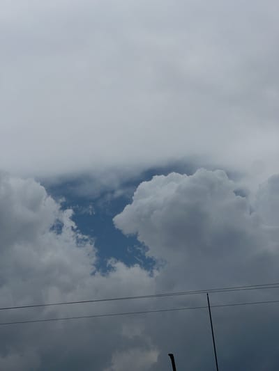 The image captures a sky scene in La Esperanza, Honduras, featuring a mix of cloud cover. A large mass of cumulus clouds dominates the center and right side, showing dark and light gray tones. Patches of blue sky are visible between the clouds. To the left, a hazy, light gray overcast obscures the sky. Power lines are visible extending from the top right corner.