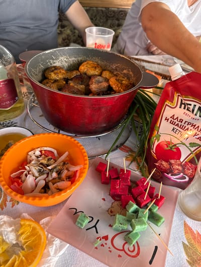 The image depicts an outdoor gathering with food and drinks, likely a picnic or barbecue, set against a rural backdrop. In the foreground, an orange bowl holds a salad composed of seafood, onions, and red peppers. Adjacent to it, a sliced orange rests on crumpled white paper. A glass plate with colorful cube-shaped appetizers, some red and some green, impaled on toothpicks, is also visible. To the right, a bottle of Heinz ketchup is prominent, alongside a bunch of green onions. In the background, a red pot filled with meat patties sits on a metal stand. The presence of people, partially visible in the upper portion of the image, suggests a social event. The overall setting appears to be casual and relaxed. The image does not contain specific visual cues that would allow for a precise location in Nizhne-Chufichevo, Russia, as such details are not discernable from the scene itself.