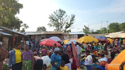 This image depicts a busy outdoor market scene in the city of Bunia, Democratic Republic of the Congo. The market is filled with numerous people, including men, women, and children, engaged in various activities such as shopping, selling, and socializing. stalls are set up with corrugated metal roofs and some are covered with colorful umbrellas, indicating an open-air marketplace. Various goods are visible, including fruits, vegetables, and packaged items. The overall atmosphere suggests a lively and bustling commercial hub.