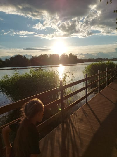 A person is sitting on a wooden boardwalk overlooking a body of water. The sun is low in the sky, casting long shadows and a warm glow across the scene, indicating it is either sunrise or sunset. The water is calm, with tall reeds growing along the bank. In the distance, there is a tree line and a hazy sky filled with clouds. The boardwalk has a rustic, wooden railing. The person is viewed from the side and appears to be looking out at the water and sky. The overall mood is peaceful and serene. The image was taken in Razvetye, Russia.