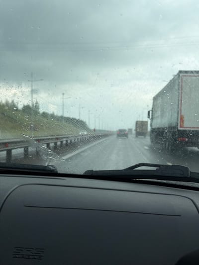 This media file depicts a view from inside a vehicle, likely a truck, driving on a highway in Устиново, Russia. The scene is captured during daylight hours, under overcast and rainy weather conditions, as evidenced by the raindrops on the windshield and the general dimness of the sky.
The primary subjects are several large trucks traveling on the highway, seen through the rain-streaked windshield. The vehicle the photo is taken from is following these trucks. In the foreground, on the right side of the frame, a portion of the vehicle's interior is visible, including the dashboard and a windshield wiper. The dashboard has an "SRS" logo embossed on it, which typically signifies an airbag system.
The activity taking place is transportation along a highway. The rain suggests a potentially hazardous driving situation, and the presence of multiple large trucks implies a commercial transport route. The setting appears to be a typical highway environment with a concrete barrier separating lanes of traffic and possibly some trees or vegetation visible beyond the barrier on the left. The overall mood conveyed is one of travel in challenging weather.