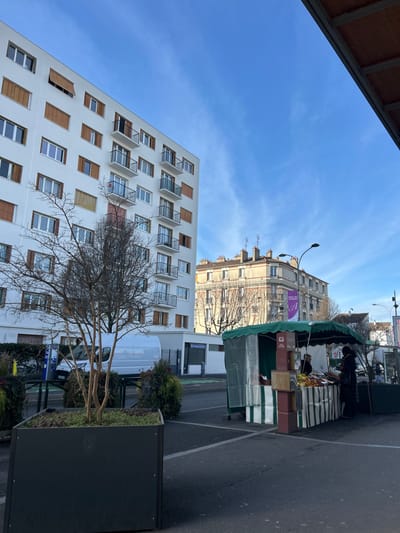 The image depicts an urban outdoor scene in Alfortville, France, under a clear blue sky with sparse cirrus clouds. In the foreground, a large, black, square planter contains a leafless deciduous tree. To the left, a modern, white multi-story residential building with numerous windows, some featuring closed brown wooden shutters, rises prominently. Multiple balconies with black metal railings are visible on this building.
Further into the scene, a white panel van is parked near the base of the modern building, partially obscured by foreground elements. In the midground, to the right, a market stall with a green and white striped awning and side panels is set up on an asphalt surface. Various produce, including fruits like oranges and apples, are displayed on tables beneath the awning, alongside stacked cardboard boxes. Two individuals are present near the stall: one person is positioned behind the stall, and another person, wearing a dark coat, stands in front of it, seemingly engaging in a transaction or selecting items.
In the background, behind the market stall, a more traditional, light-colored building with multiple floors, a mansard roof, and visible chimneys is situated. A purple banner with a white 'A' logo is affixed to the side of this building. Streetlights are also visible within the scene. A brown, sloped roof or awning structure occupies the upper right corner, suggesting the vantage point is from beneath a similar overhead structure. The lighting indicates daytime conditions.