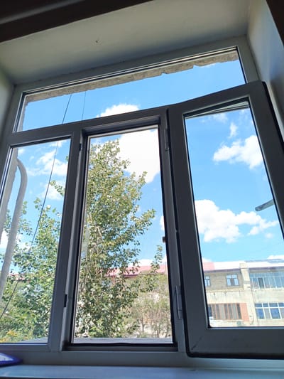 The image is a close-up, interior view of an open window, looking outwards. The window frame is white and appears to be made of vinyl or a similar material. The window itself is comprised of multiple panes, with some panes open, allowing a view of the exterior.

Outside the window, the sky is a clear blue with scattered white cumulus clouds. A large tree with green foliage is visible in the foreground, partially obscuring the view of buildings behind it. The buildings appear to be multi-story structures with a mix of architectural styles, suggesting an urban or semi-urban environment. One building has a red-tiled roof.

The scene is consistent with a residential or commercial area in Nukus, Uzbekistan, given the characteristic architecture and climate. There are no people visible in the image, and the focus is on the window and the view it provides. The lighting suggests it is daytime, likely late morning or early afternoon due to the brightness and the position of the sun, casting subtle shadows. A windowsill is visible at the bottom of the frame, with a slight reflection or glare on its surface. There are some dark marks or smudges on the window frame and the interior wall above the window.