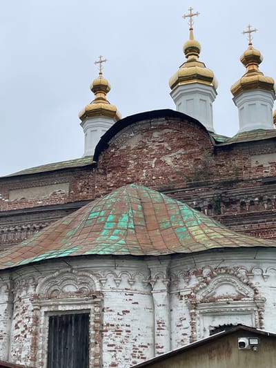 The image displays a section of a Russian Orthodox church in Kstynino, Russia. The foreground features a weathered, multi-faceted roof covered in oxidized copper, showing patches of green patina over a reddish-brown base. Behind this, the church's facade is visible, constructed from aged red brick with significant signs of deterioration, including crumbling mortar and exposed brickwork. Decorative elements, likely plasterwork, are present around arched windows and cornices, but these also show wear and tear, with some sections appearing damaged or missing. Above the brickwork, several gilded onion domes topped with crosses pierce the overcast sky. The domes are brightly polished and contrast sharply with the weathered architecture below. The overall impression is one of historical significance and enduring, yet deteriorating, beauty.